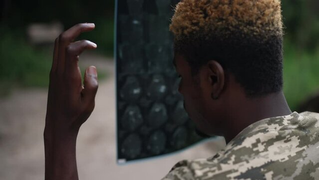 Zoom in to sad frustrated African American soldier in wheelchair with X-ray sitting in summer park thinking. Shooting over shoulder of young man with paralysis procrastinating outdoors. Slow motion