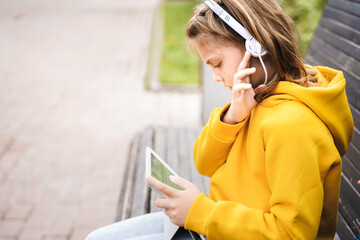 Girl in headphones, headset with tablet. Study, online conference, lesson, education. Remote learning on street bench in school yard, park. Schoolgirl with backpack. Chat, talk with friends, teacher