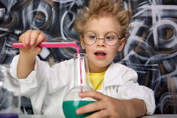 Cute scientist with glasses mixes reagents in lab. Beautiful little boy scientist is intrigued and excited, exploring colored chemical liquids in the laboratory using test tubes of different sizes.