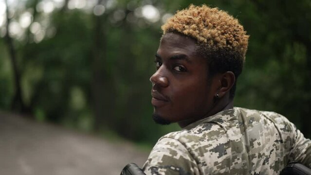 Close-up African American young man in wheelchair turning looking at camera smiling. Portrait of confident positive handsome soldier in military T-shirt posing in summer park in slow motion