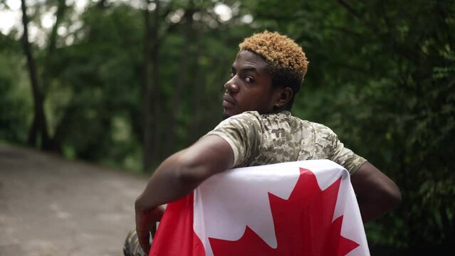 Live camera zoom in to confident positive African American man in wheelchair turning posing in slow motion in park. Young handsome soldier looking at camera smiling with Canadian flag on device