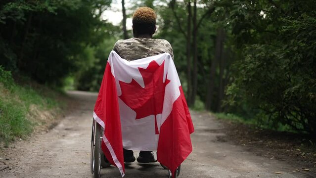 Back view wide shot wheelchair with Canadian flag and African American young man thinking admiring nature. Unrecognizable soldier procrastinating outdoors in park. Slow motion