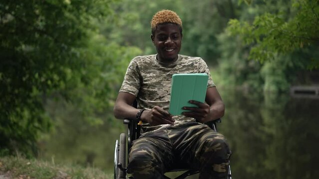 African American smiling soldier waving greeting at video chat on tablet sitting in wheelchair on lake shore. Portrait of positive inspired young handsome man messaging enjoying tourism in forest
