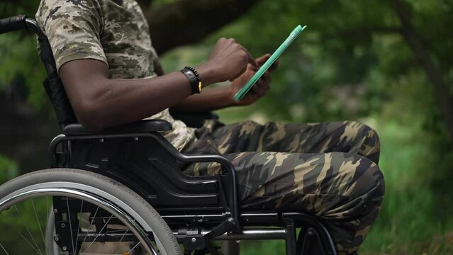 Side view unrecognizable African American man in wheelchair messaging online on tablet outdoors in slow motion. Young brave soldier surfing Internet resting in green summer park