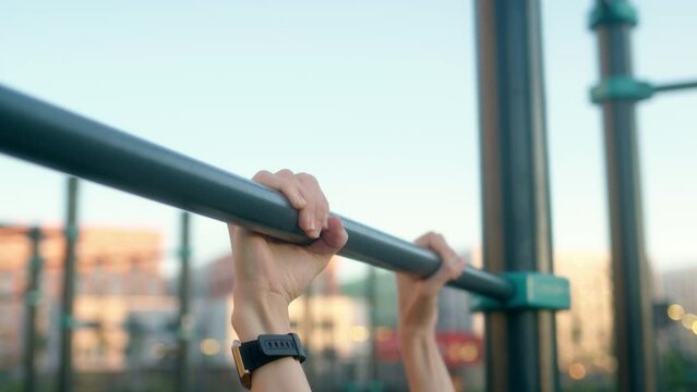 young woman in morning practices pull-ups on bar in black hoodie with pigtail. attractive brunette engaged in workout on sports ground in courtyard of house. close up view of hands pulling up.
