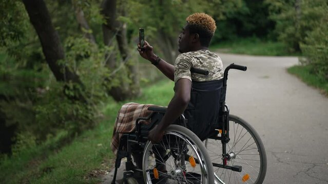 Curios African American man in wheelchair taking photo of nature on smartphone in summer forest. Side view wide shot of young military tourist enjoying travelling in park outdoors. Slow motion