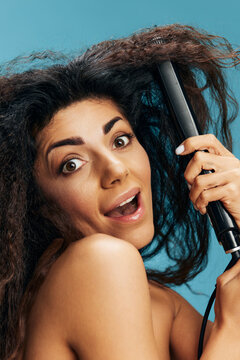 Excited pretty Latin curly woman using hair straightener, looking aside, posing isolated on blue wall background. Hair routine concept, haircare, dry damaged hair ironing, hairdressing