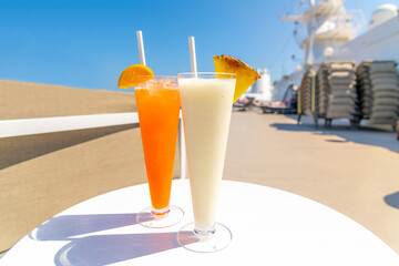 Two colorful mixed drinks with fresh fruit and a straw on the upper deck of a luxury cruise ship at sea on a sunny day.