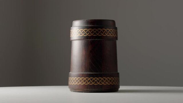 Wooden oak mug with a handle is spinning on white table. Brown mug with ornament isolated on dark gray studio background. Handmade wooden tankard for beer or kvass. Rustic background.