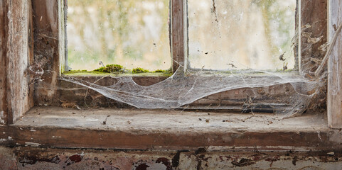 Abandoned, dirty and dusty window covered in spiderwebs in empty house from poverty and economic crisis. Old, damaged and weathered wooden windowsill and wood frame rotting from dampness and neglect