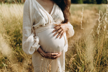 a young woman in a white linen dress wrapped her arms around her pregnant belly. body of pregnant...
