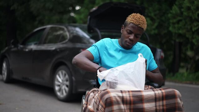 African American young man in wheelchair with groceries outdoors with blurred black car at background. Portrait of sad young guy waiting for driver shopping. Slow motion