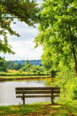 Fototapeta na wymiar Park bench overlooking the view by the lake in the countryside. Scenic view of trees and greenery by a river with an empty public bench in summer. Landscape view of an idyllic natural environment
