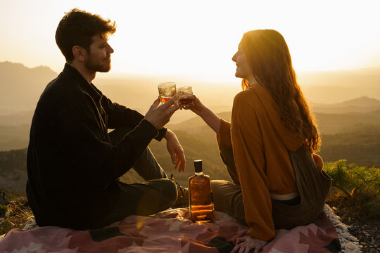 Cute couple having a drink making a toast on top of mountain at sunset