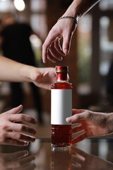 Glass bottle with alcohol drink with mockup on mirror table. Male people's hands reach for the bottle. Concept of celebration holiday in pub