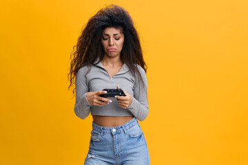 Nervous young latin woman 20s in casual top holding and playing pc games with console joystick, biting lips isolated on orange yellow background studio portrait. People lifestyle concept.