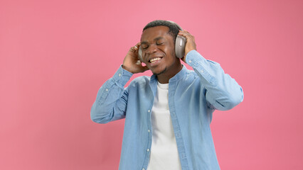 Portrait dancing black man on headphones. Slow motion young happy teenager 20 years old African American in denim shirt dancing listening to music with headphones on head on pink studio background.