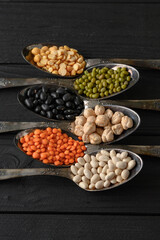 Variety of legumes in old silver spoons on a black wooden background.
