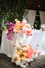 Wedding table decor with flowers for the newlyweds. On the corner of the table is a bouquet of yellow gerberas, white chrysanthemums, and pink roses. White tablecloth and candles in the background.