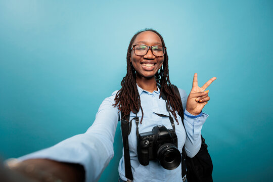 Excited joyful photography entusiast taking selfie photos while standing on blue background. Happy smiling photographer with DSLR camera giving peace sign to audience while recording daily vlog video.