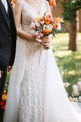 The bride in a shiny white dress with a pattern holds a wedding bouquet of white, yellow, orange, and pink roses and peonies.