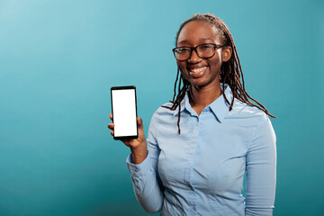 Cheerful smiling young woman holding modern smartphone device with isolated mockup screen on blue background. Pleased and confident adult person having phone device with blank copyspace template.