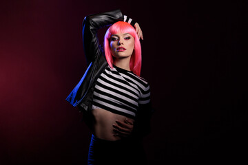 Portrait of female punk rocker posing in dark shadows, feeling attractive and confident in studio. Young woman with pink hair and stylish fashion, wearing biker jacket over colorful background.