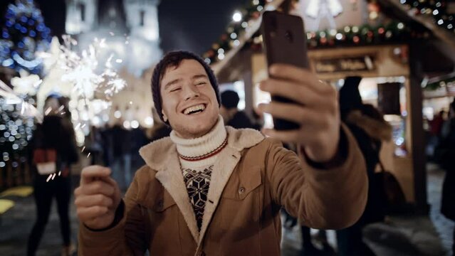 Beauty tourist man smiling happy city street looking at mobile phone screen and congratulate on holiday via video chat friends with falling sparkling glowing sparklers fireworks light in europe night.