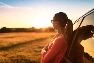Female standing next to car looking out to an open field at sunset. On a road trip adventure, Feeling at peace in nature concept.	