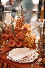 Wedding banquet table is decor with orange flowers and burning candles on an orange tablecloth. The restaurant menu is on a plate with a silk napkin.