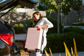 Excited woman talking on phone while loading suitcase into car trunk