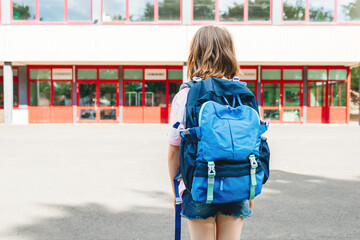 A teenage girl with a school backpack on her back stands in front of the school and looks at her. Beginning of the school year for students. Back to school