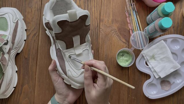 Girl's hands paint sneakers with brush on wooden table. Designer's workshop. Palette with brown and green acrylic and other tools on desktop. Hobbies, creativity and handmade work at home.