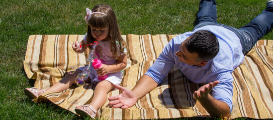 Image of a young dad sitting in a green meadow playing with his daughter making soap bubbles. Reference to the bond between father and daughter. Horizontal banner