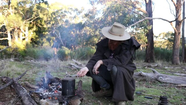 A bushman in an oil skin stockman jacket sits around a fire with his billy boiling in the high country.