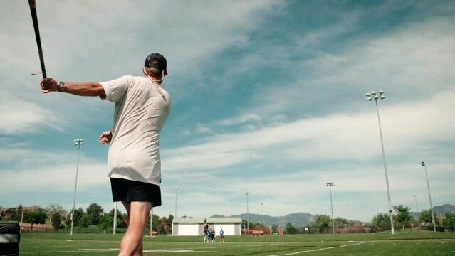 A low wide shot of a professional male baseball player practices tossing a ball into the air and hitting it.  A dramatic sky frames his athletic form as he practices hitting balls for others to catch.