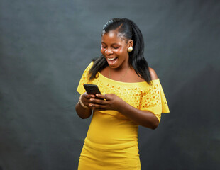 A happy African Nigerian female lady in yellow dress joyfully using, typing and looking at the smart phone in her hands