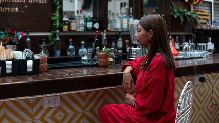 Latin girl in a red dress sitting in front of a bar counter. Party girl in a night club. Girl ordering a drink in a bar. Hispanic woman in a bar waiting for her order.