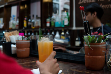 Hand of a girl in a red dress holding a glass of juice. Unrecognizable girl at a bar counter with the waiter in front of her. Wooden bar with rustic aesthetics of a restaurant.
