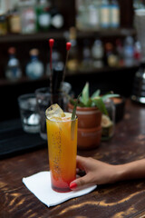 A finely prepared drink with a napkin underneath on a wooden table being taken by a girl's hand. A variety of alcoholic beverages are seen in the background. Yellow drink on a dark background.