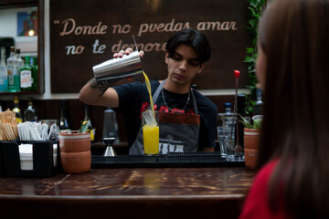 Bartender at the bar counter preparing a drink in front of a client dressed in red with brown hair. Waiter preparing a yellow drink. Girl in a nightclub in front of the bartender.