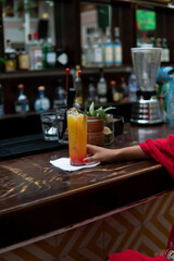 A yellow drink on a wooden table and the hand of an unrecognizable girl in a dress with red nails. A glass of juice on a bar counter. In the background there is a wide variety of bottles.