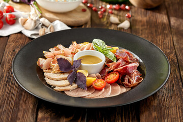 Meat platter on black plate on wooden background. Meat assorted with sauce and basil. Composition with Italian antipasti in rustic style. Meat delicatessen on dark wooden table with ingredients.
