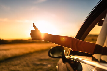 Getting away from it all. Happy person woman on a road trip with thumbs up arms out of the car facing a beautiful sunset.  - obrazy, fototapety, plakaty