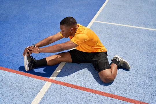 Athletic young black guy stretching legs at stadium