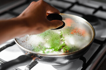Close-up of human hand opening frying pan with food on gas stove. Selective focus on misted glass lid of frying pan