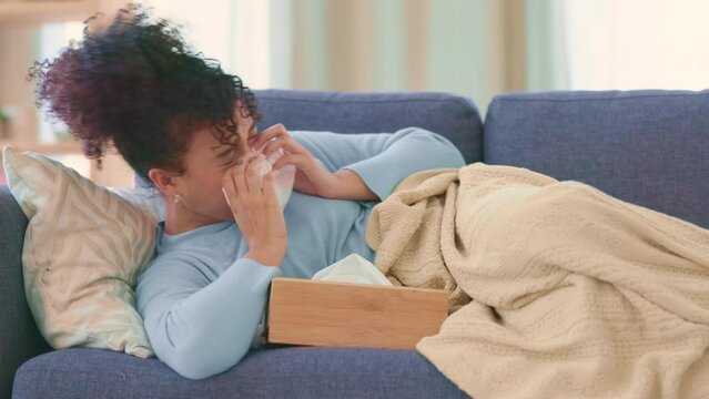Woman sick with the flu on a couch at home, sneezing and blowing nose. Young female suffering from sinus, allergy or Covid19 while isolating. Lady taking off from work to recover from seasonal cold