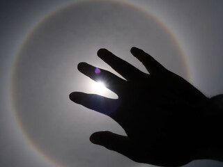 Beautiful photograph of the sun with a circular rainbow surrounded by a bright sky and white clouds with shadows of hands reaching out. Phenomenon, sun halo.
