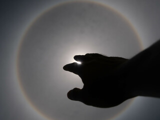 Beautiful photograph of the sun with a circular rainbow surrounded by a bright sky and white clouds with shadows of hands reaching out. Phenomenon, sun halo.