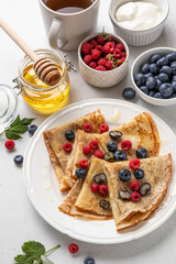 French crepes or traditional Russian blini on white plate on light background. Thin pancakes served with raspberries, blueberries, tea, honey and whipped cream. Summer concept. Selective focus.